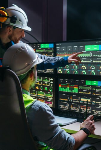 Head engineer and factory operator wearing safety vests and hard hats wollowing product process on factory uses SCADA system and industry 4.0. Two operators controll assembly line using screens with human-machine interface Head engineer and factory operator wearing safety vests and hard hats wollowing product process on factory uses SCADA system and industry 4.0. Two operators controll assembly line using screens with human-machine interface